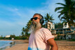 © artiemedvedev - Grey-bearded senior man enjoying music on tropical beach. Stylish elder in sunglasses and wireless headphones. Relaxing, having fun by sea waves under palm trees. Elegant modern lifestyle.