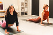 © Studio Marmellata - Two women perform the upward-facing dog yoga pose on black mats in a well-lit studio with shelves and artwork in the background, wearing athletic clothing and focusing on their practice