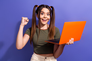 Wall Mural - Excited young woman holding a laptop, cheering with joy, and standing against a vibrant purple background