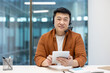© Liubomir - Portrait of successful Asian worker with headset and tablet computer in hands. Man smiling and looking at camera, working inside office at workplace. Online support service worker.