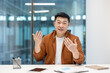 © Liubomir - Webcam view, video call, Asian man looking at camera, smiling and communicating with colleagues and partners remotely. Successful businessman working inside office at workplace.