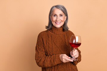 Wall Mural - Smiling elderly woman holding wine glass against beige background, dressed casually in brown sweater