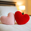 © homydesign - Two heart-shaped pillows, one pink and one red, resting on a white bed with a softly blurred background featuring a bed and a lamp