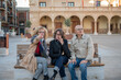 © C&A - Three middle-aged friends sitting on a bench in a historic plaza, one pointing at something in the distance while enjoying a casual conversation