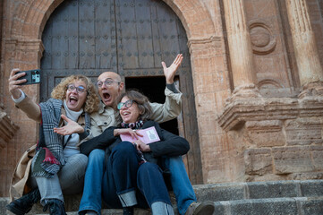 Three senior friends taking a joyful selfie on historic stone steps in front of a grand wooden door, in a small town in Spain