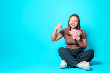 A cheerful woman seated cross-legged against a vibrant background, displaying cash and approval. This image perfectly represents financial success, happiness, and positive emotions.