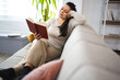 © Jelena - Woman reading a book while relaxing on a sofa at home
