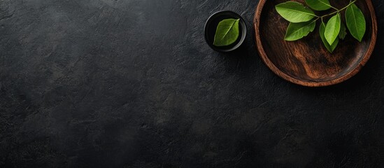 Poster - Black stone surface with green leaves in a wooden bowl and small black dish Copy Space
