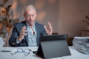 Wall Mural - Senior businessman sitting at his desk in office at night having a video conference on a tablet looking surprised and gesturing with his hands