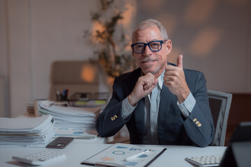 Wall Mural - Satisfied senior manager showing thumbs up gesture while sitting at desk in office late at night, working with financial reports, smiling at camera