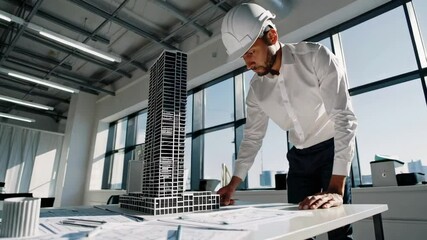 Poster - Professional architect wearing hardhat meticulously examines a scale model of a skyscraper, carefully reviewing blueprints laid out on a table in a contemporary, light filled office space