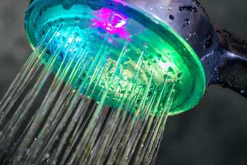 A close-up of a shower head illuminated with vibrant LED colors, emitting flowing water streams. The droplets and colors create a modern and vibrant design.