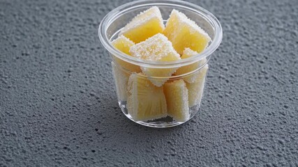 Poster - A minimalist composition where a single clear tub of frozen pineapple chunks sits on a textured surface, emphasizing the icy coating on each piece.