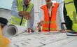 © wichayada - Construction team pointing at project plans and discussing layout details in a professional office setting