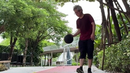 Wall Mural - A senior East Asian man is practicing and playing basketball alone in a public park.