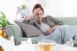 © amedeoemaja - Young woman lies on the couch, holding her head and stomach in pain, suffering from a headache and abdominal discomfort, due to period cramps or flu, trying to relieve with tea and medication pills