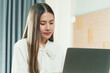 © Ekkasit A Siam - Asian woman in white blouse working on laptop at home, appearing focused. Modern home interior with wooden headboard and soft lighting create a comfortable workspace for remote productivity.
