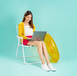 © sitthiphong - Happy smiling young Asian woman in summer sit in deckchair and type text on a laptop in blue background.