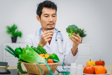 Wall Mural - An Asian male nutritionist with a beard, wearing a lab coat, sits at a desk for an online consultation. His workspace includes fruits, vegetables, laptop, health supplements, promoting wellness.