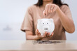 © Phushutter - A young Asian woman in a light brown t-shirt, sitting with a piggy bank, coin, calculator, mobile, and pen. She focuses on financial stability, saving for emergencies, education, and future goals.