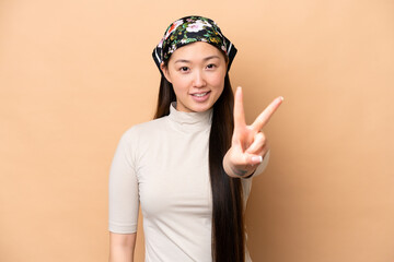 Wall Mural - Young Chinese woman isolated on beige background smiling and showing victory sign