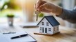 © felix_brönnimann - Hand holds house key above miniature model home on desk, symbolizing property ownership, real estate investment, or home buying process for first-time buyers.