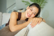 © wattana - Beautiful young woman relaxing on a couch in cozy living room and smiling warmly at the camera.