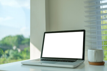 Fototapete - Laptop with a blank display is placed on a white desk near a large window with blinds.