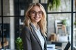© El Media - A photo of an elegant woman with blonde hair and glasses, smiling confidently at the camera while standing in front of her desk filled with office supplies like paper.