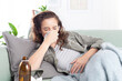 © amedeoemaja - Young woman with the flu lying on the couch, blowing her nose due to a cold and cough, suffering from breathing problem and feeling unwell, while treating herself with rest and medication