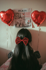 Wall Mural - A photo of a wall with red heart-shaped balloons, an open magazine, and a girl lying in bed with long, dark brown hair tied in two braids, wearing a fabric bow around her neck, with the word 