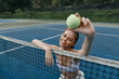 © SHOTPRIME STUDIO - Happy young female tennis player in sportswear holding a tennis ball at the net on a sunny day, showcasing energy and excitement on a blue court