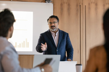 Wall Mural - Serious confident mature Indian business teacher giving seminar to students, speaking at tribune and whiteboard, explaining lecture. Senior boss talking to employees on presentation meeting