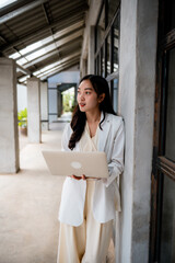 Poster - Young asian businesswoman is holding a laptop and looking away while leaning against a wall in a modern office building