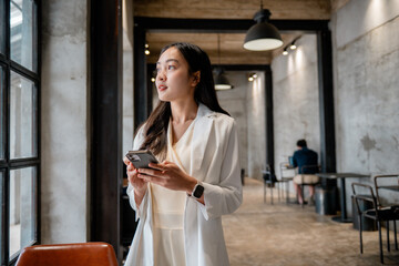 Poster - Young asian businesswoman is using smartphone and looking through window in modern office