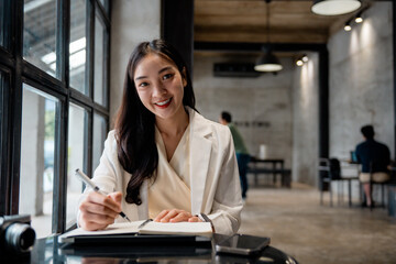 Poster - Young asian businesswoman smiling and writing on notebook in a modern co-working space