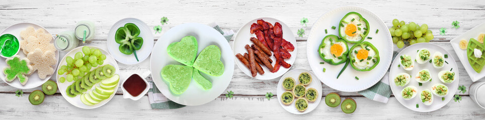 Poster - St Patricks Day breakfast or brunch table scene against a white wood banner background. Selection of green and shamrock themed pancakes, toasts, eggs, fruits and meats. Above view.