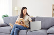 © Studio Romantic - Happy woman watching online laptop, happy girl holding cute pet puppy at home, enjoy time together with small pedigreed dog on sofa, searching for vet information, friends in internet social network