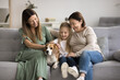 © fizkes - Three generations of women spend time at home. Cute preschooler girl stroking adorable beagle dog, resting on cozy sofa with loving young mother and older grandmother. Multigenerational family leisure