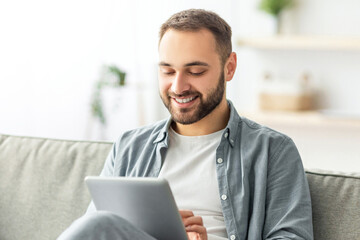 Poster - Joyful millennial man sitting on sofa with tablet computer, working or studying online at home. Happy Caucasian male participating in web lecture, taking part in webinar or video conference