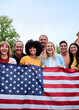© CarlosBarquero - Vertical. Multiracial group of friends holding the American flag with pride, celebrating Independence Day outdoors. Joyful atmosphere with a sense of unity and patriotism in a vibrant summer setting