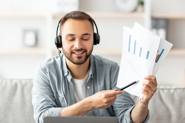 Poster - Cheery young guy in headset making video call, showing documents at webcam, using laptop, working from home. Millennial man communicating online, participating in web conference or business meeting