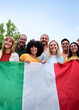 © CarlosBarquero - Vertical. Multiracial group of friends holding the Italian flag with pride, celebrating patriotism and love for sports, football. Cheerful atmosphere outdoors, unity, enthusiasm, and cultural pride