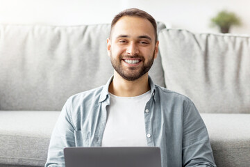 Poster - Full length of happy young guy sitting cross legged on floor, using laptop computer, smiling at camera indoors. Positive millennial freelancer working online from home, attending business webinar