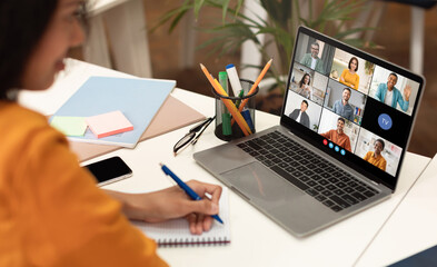 Poster - A woman sits at a desk with a laptop open, displaying a video conference meeting with several participants. She is taking notes in a notebook with a blue pen, her focus on the screen.