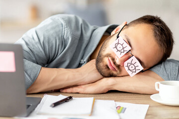 Poster - Lazy unproductive young guy wearing funny sticky notes with open eyes on his glasses, sleeping at workplace. Tired overworking millennial man falling asleep at home office