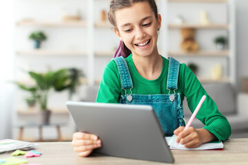 Poster - Remote Education Concept. Smiling young teenage girl sitting at desk, holding and using digital tablet writing in her notebook. Schoolgirl watching online course, taking notes, doing homework