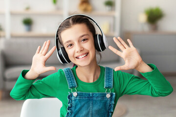 Poster - Portrait of smiling girl wearing wireless headphones talking and waving two hands, looking at camera, showing ten fingers, webcam point of view. Teen making video call with teacher or recording vlog