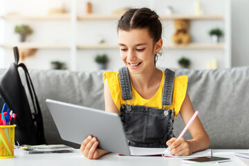 Poster - Children and Education Concept. Portrait of positive teen girl using doing homework writing in notebook sitting on couch at desk, holding and using tablet pad computer, enjoying distance homeschooling