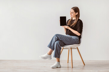 Poster - Charming young woman showing tablet with empty screen, sitting on chair against white studio wall, mockup for website or new app design. Millennial lady demonstrating touch pad display template